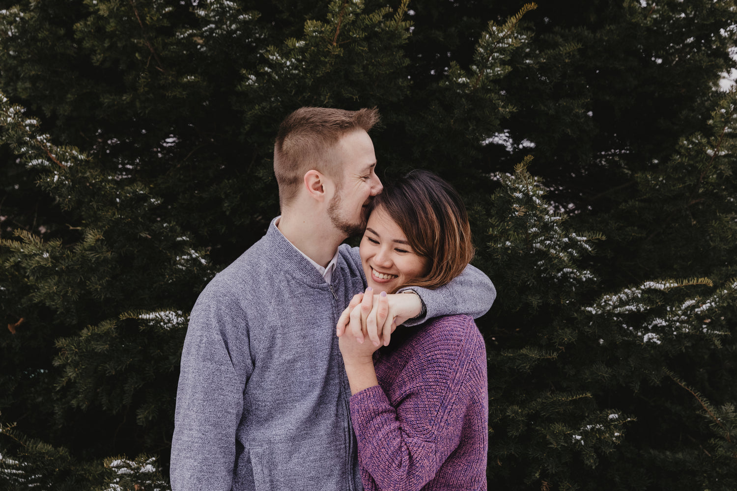 A couple in front of a tree holding hands while the man kisses the woman's head